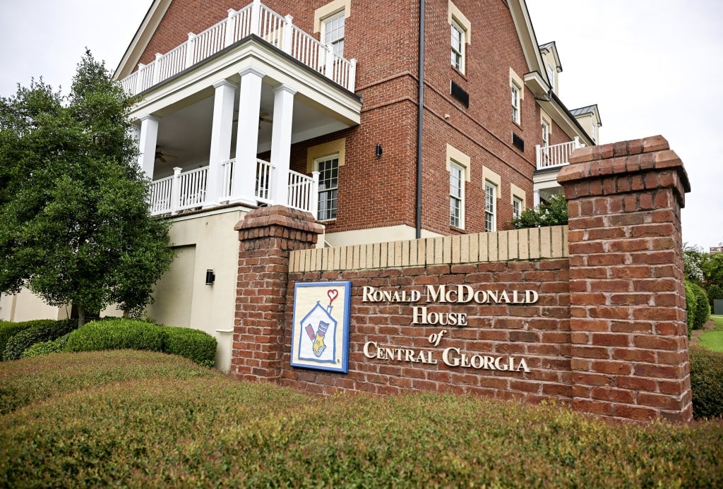 The image shows the exterior of a brick building with a white balcony and a sign that reads "Ronald McDonald House of Central Georgia" on a brick wall in the foreground. There are bushes and a tree in front of the building. The sign includes a small illustration of a house with a heart and a helping hand.