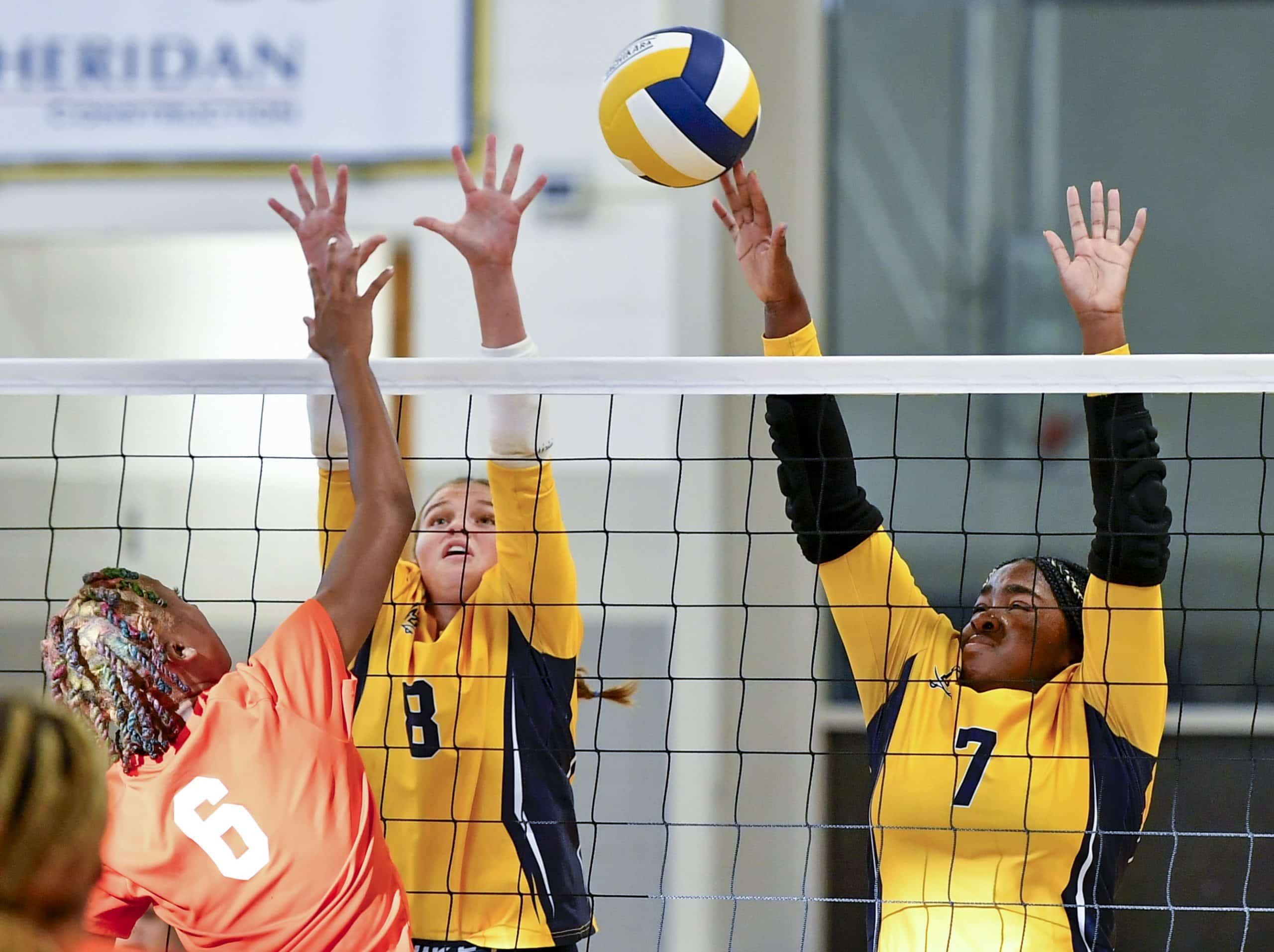A volleyball player in an orange jersey is spiking the ball over the net while two players in yellow jerseys jump to block it.