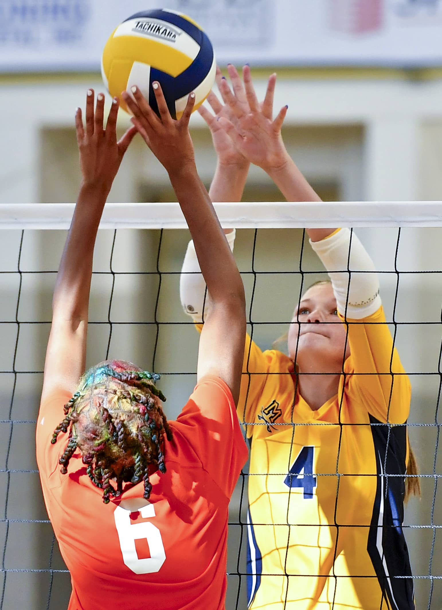 Two volleyball players one in an orange jersey and one in yellow jersey battle for the ball over the net.