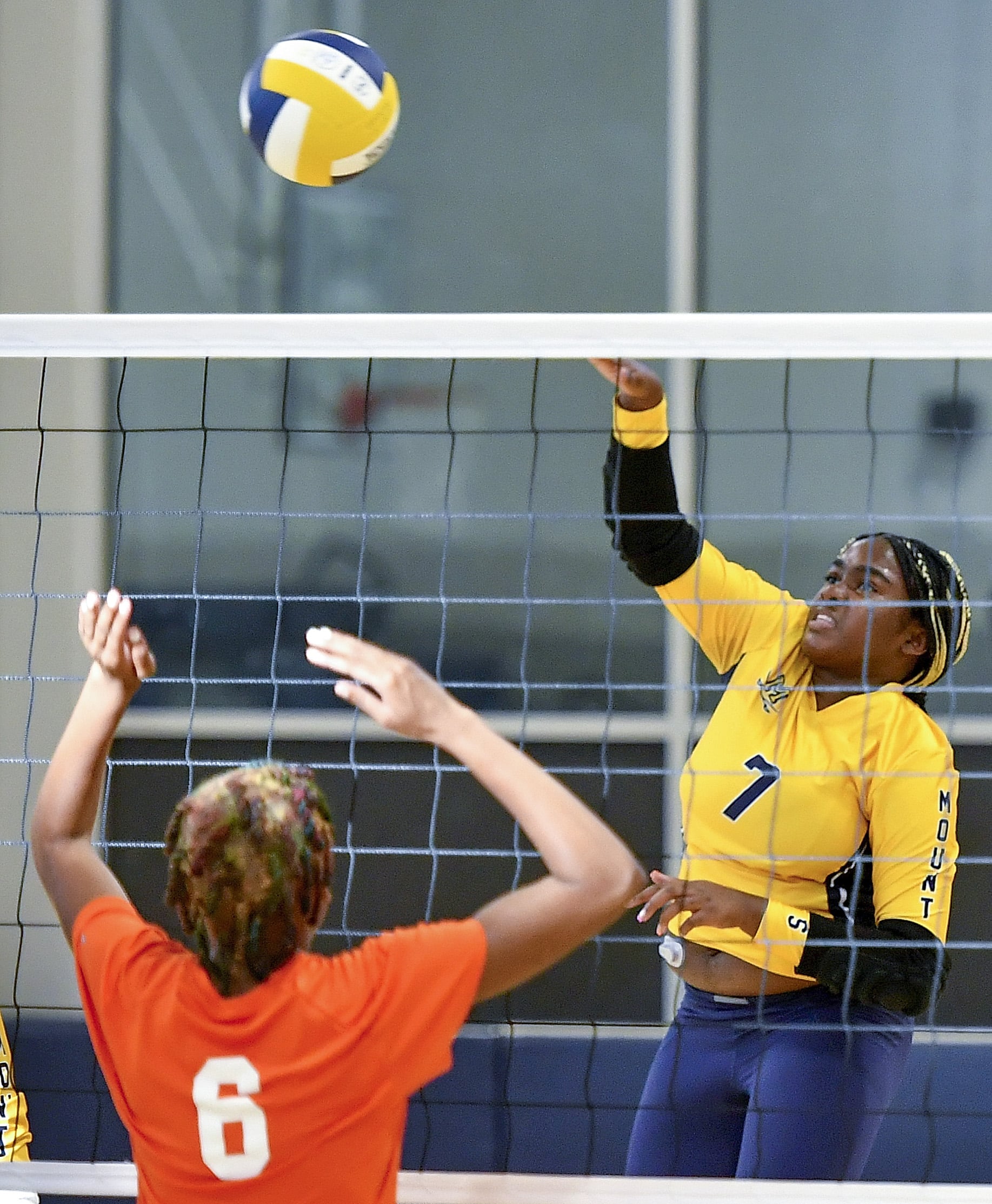 A volleyball player in a yellow jersey is jumping to spike the ball while two players in orange jerseys jump to block her.