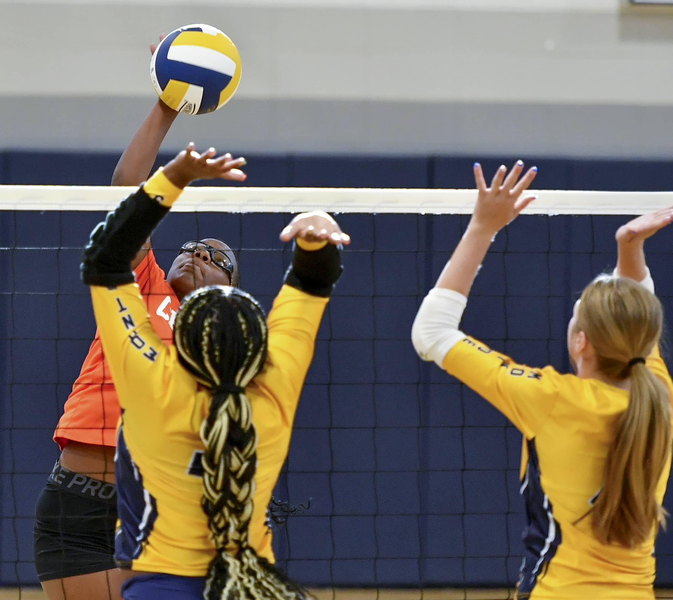 A volleyball player in an orange jersey is spiking the ball over the net while two players in yellow jerseys jump to block it.