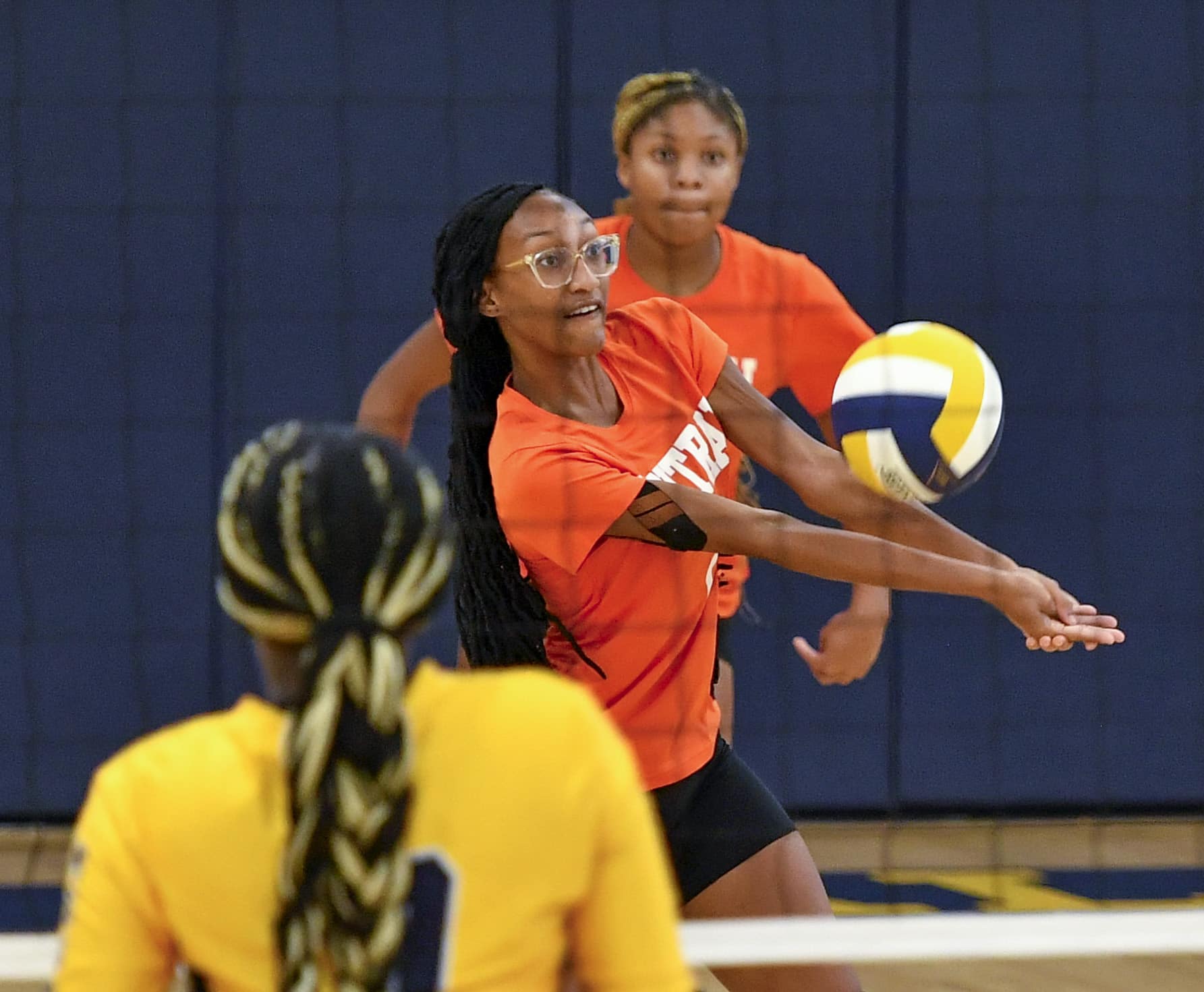 A volleyball player in an orange jersey sets the ball while a teammate looks on from behind.