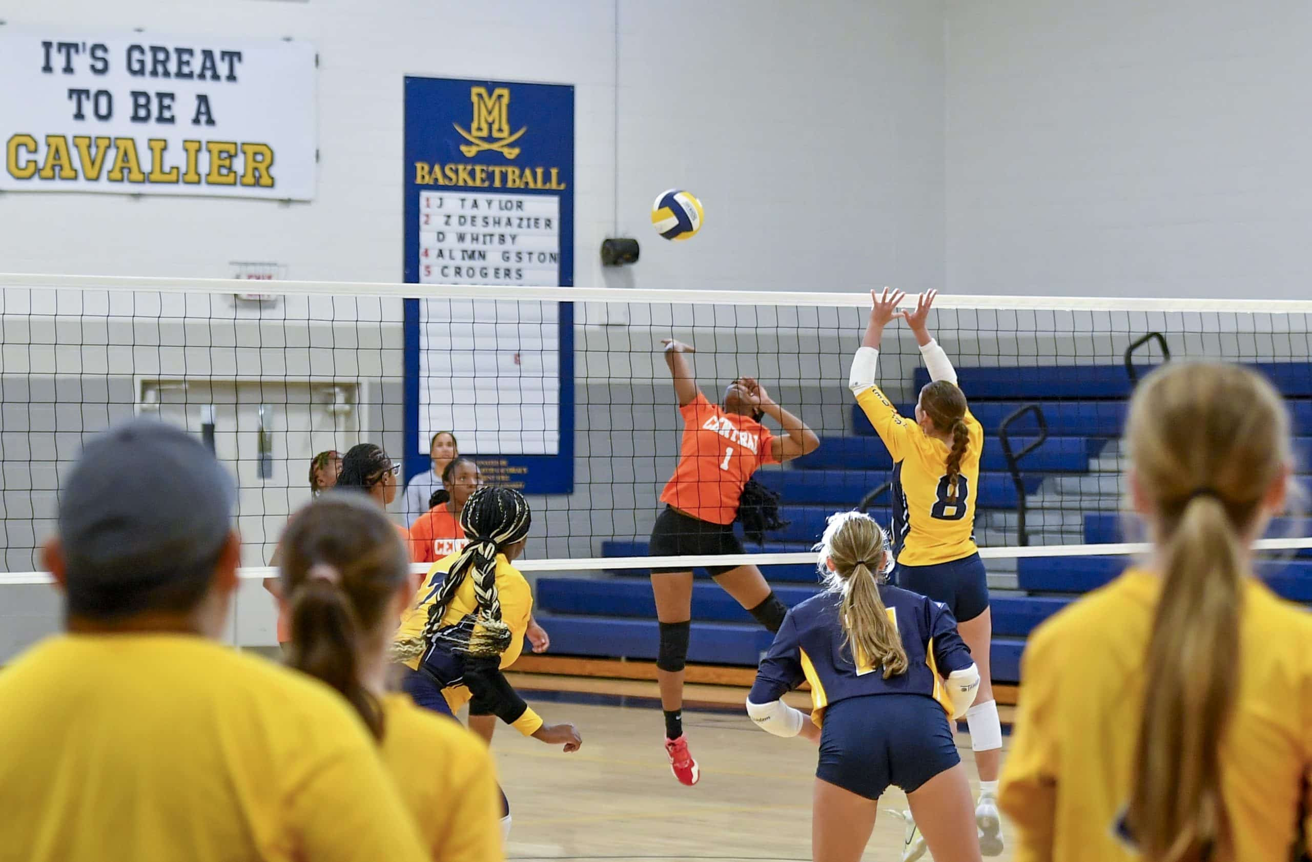 A volleyball player in an orange jersey is spiking the ball over the net while two players in yellow jerseys jump to block it.