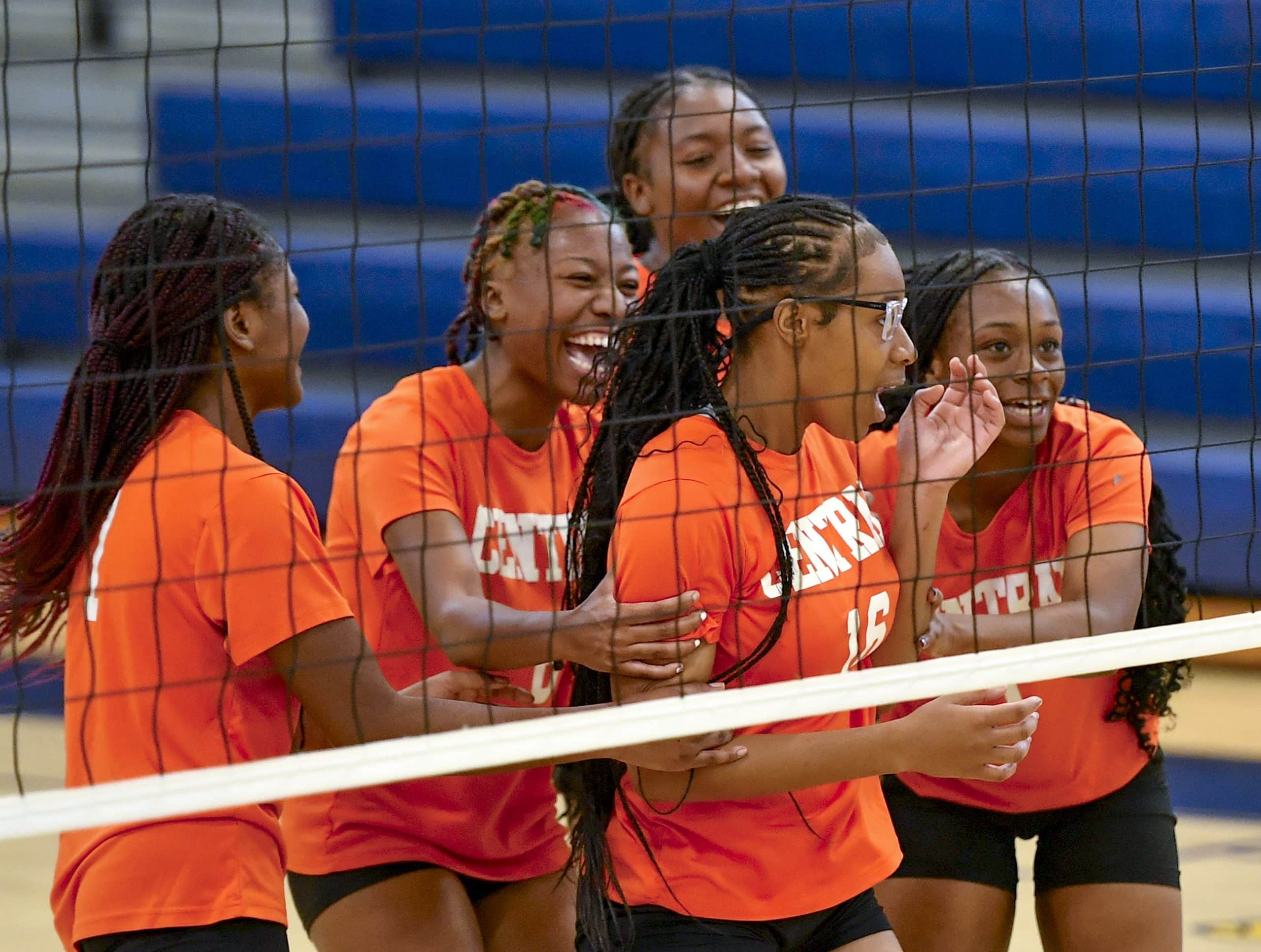 A volleyball player in an orange jersey is greeted by teammates near the net.