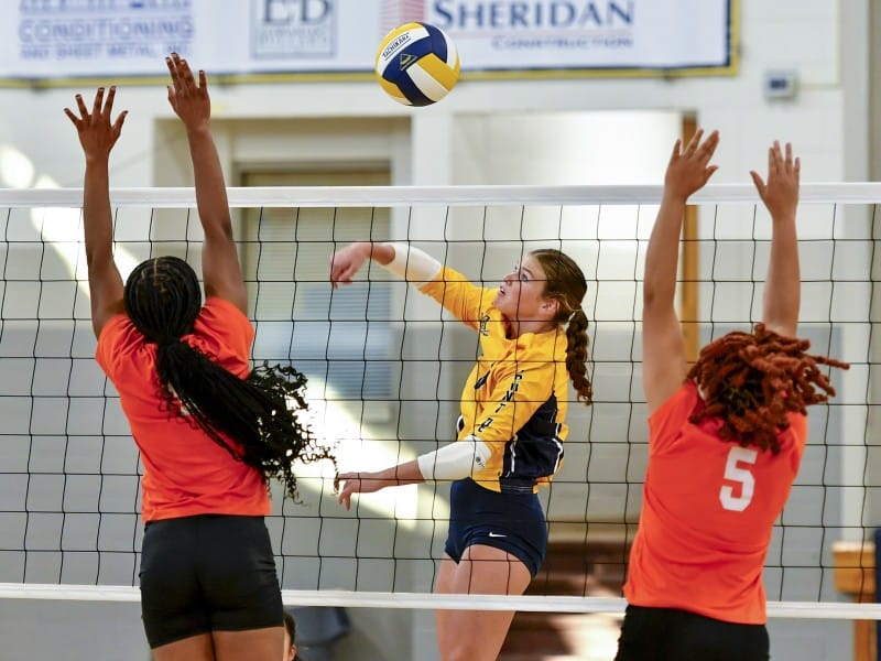 A volleyball player in a yellow jersey is jumping to spike the ball while two players in orange jerseys jump to block her.