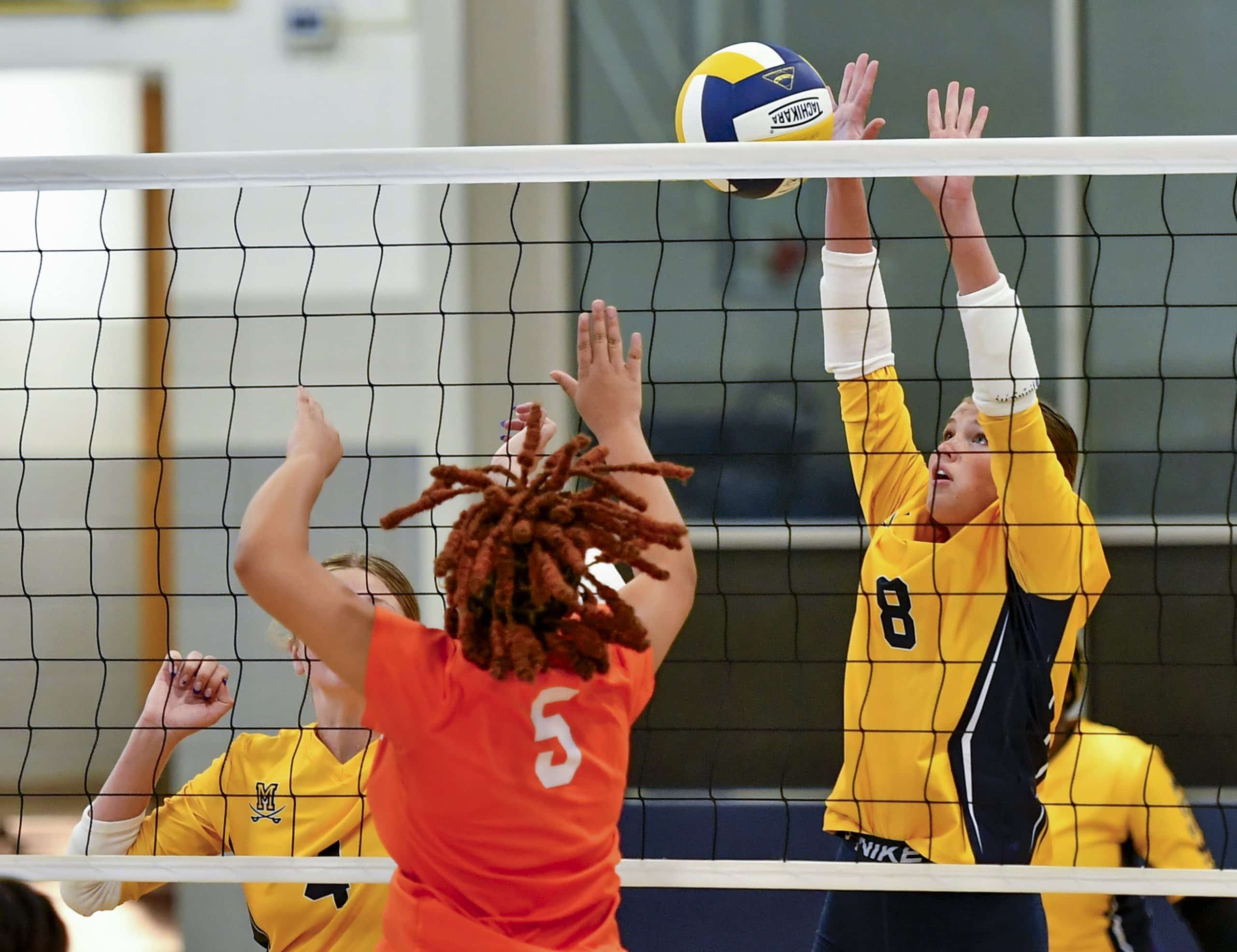 A volleyball player in a yellow jersey is jumping to block the ball after a player in an orange jersey spikes the ball.