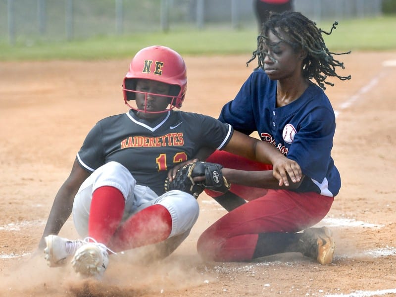 A player in a black jersey, grey pants and red helmet slides in a cloud of dirt as a player in a blue jersey with red pants tries to tag her with the ball in her glove.