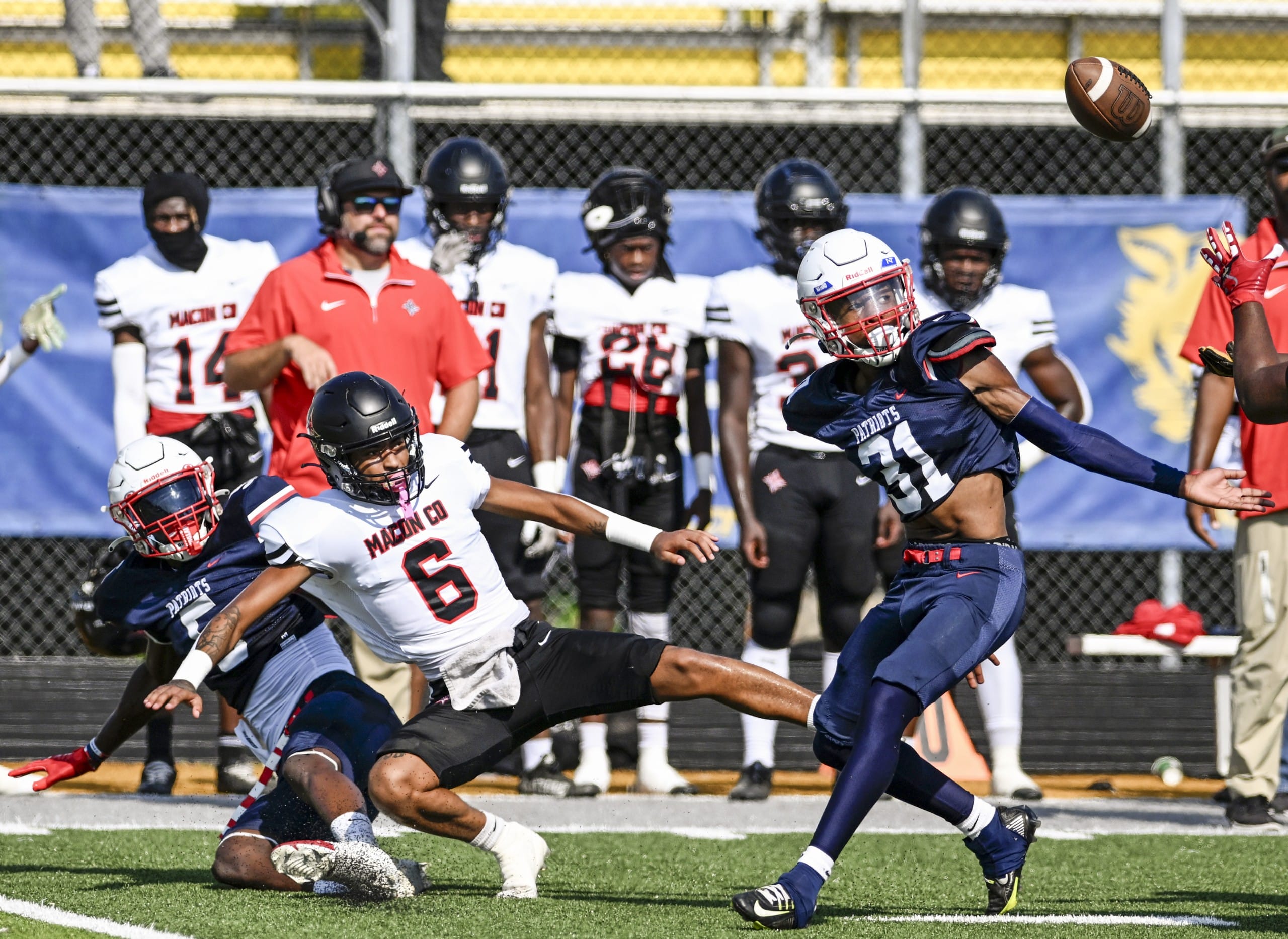 A football player wearing a blue uniform stretches for a ball in the air behind him while being a player in a white uniform with the number 6 falls to the ground.