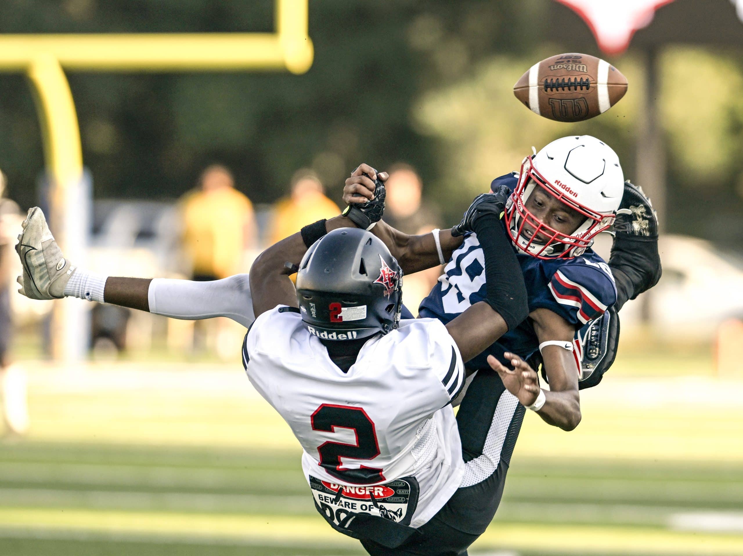 A football player wearing a blue uniform with the number 18 is tackled to the ground with the ball in the air by a player in a white uniform with the number 2.