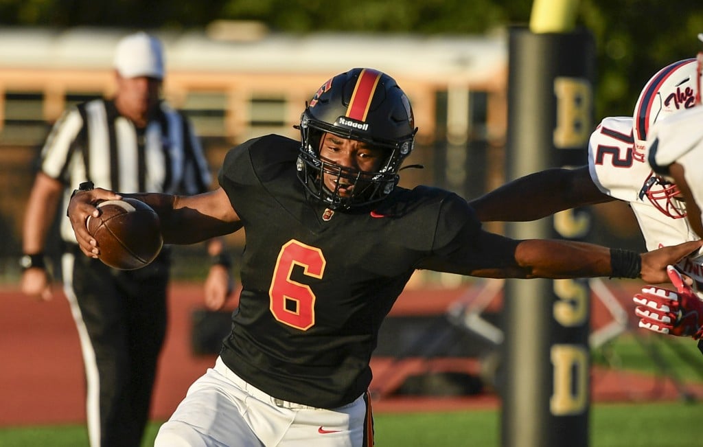 A football player in a black jersey with a red number, a black helmet with red stripes and white pants holds a football in his right hand while stiff-arming another player with his left.