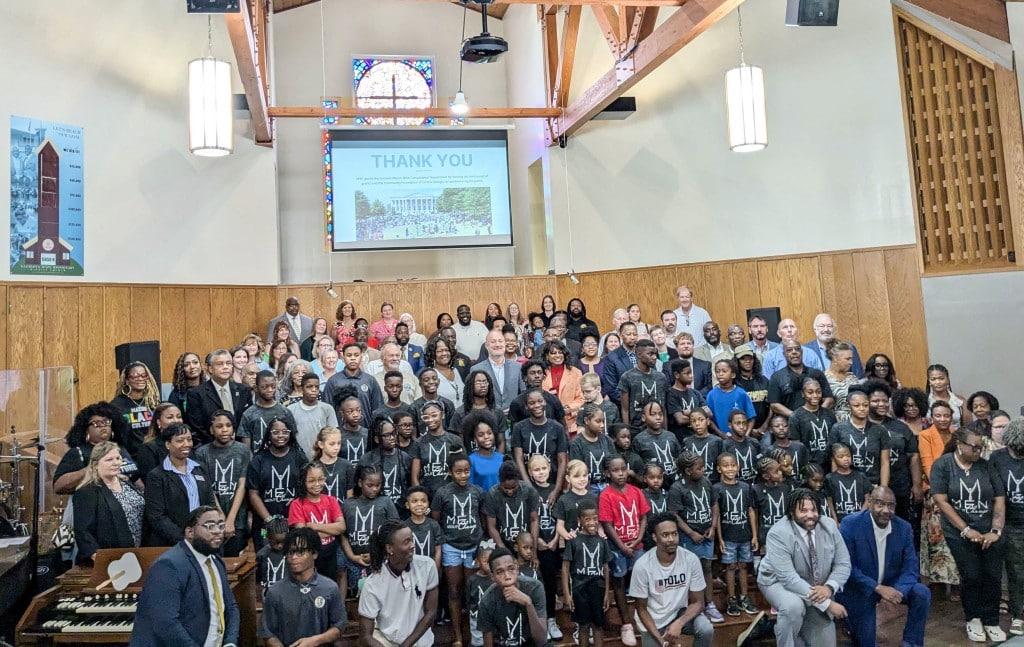 A large group of people, including many children and adults, pose together inside a church. Most of the children are wearing matching black shirts with white designs. A screen behind them displays the word "THANK YOU" above a photograph of a large gathering outdoors. The setting features wooden walls and a stained-glass window.