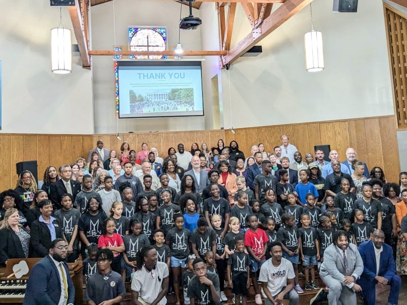 A large group of people, including many children and adults, pose together inside a church. Most of the children are wearing matching black shirts with white designs. A screen behind them displays the word "THANK YOU" above a photograph of a large gathering outdoors. The setting features wooden walls and a stained-glass window.