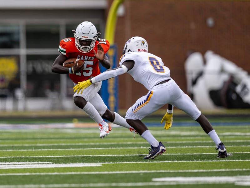 A player in an orange jersey, white pants and white helmet runs sideways while toting the football in attempt to avoid a defender wearing all-white with a #8 on his jersey.