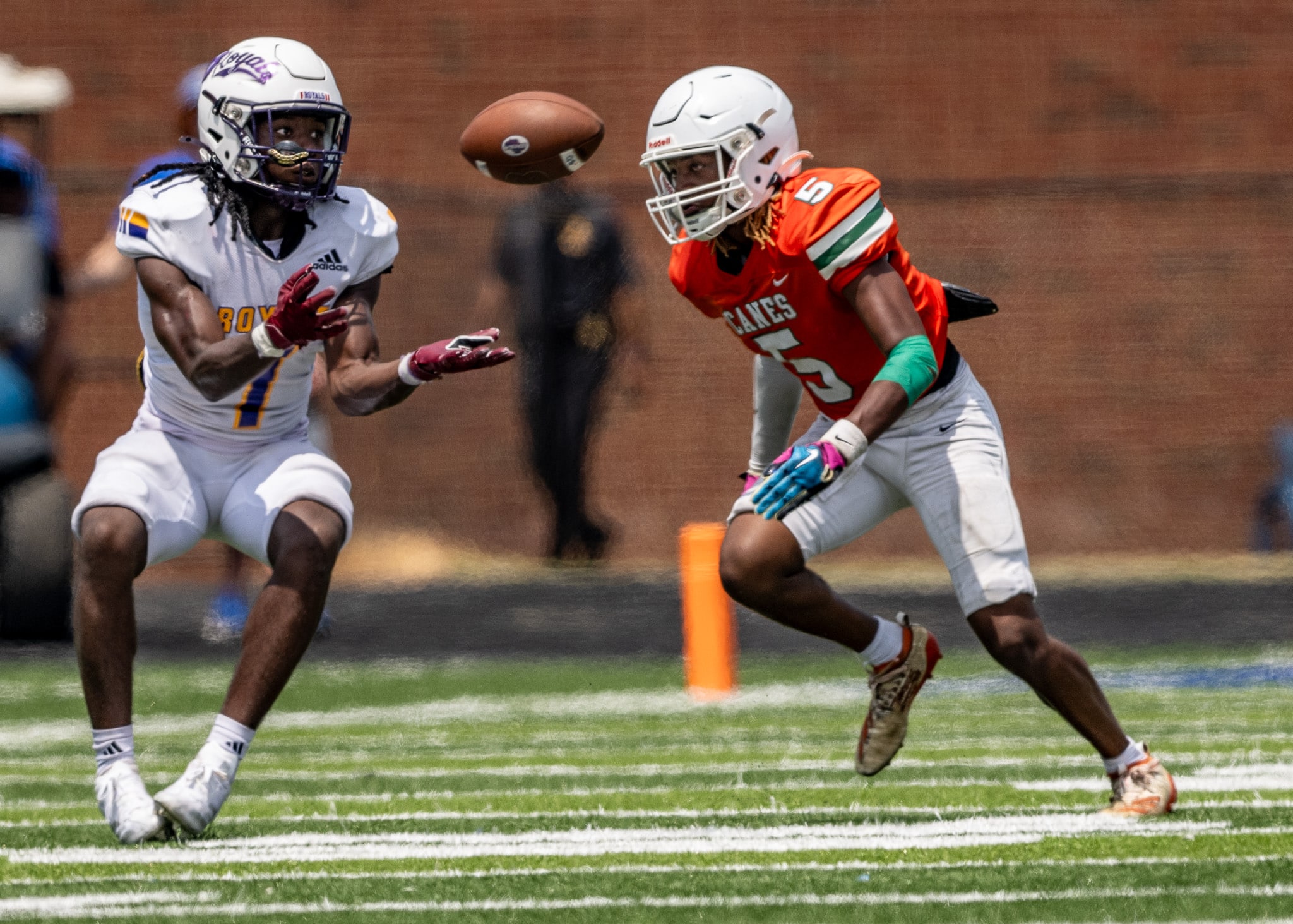 A football player wearing a white uniform with the number 7 waits for a thrown ball while being defended by a player in an orange uniform.