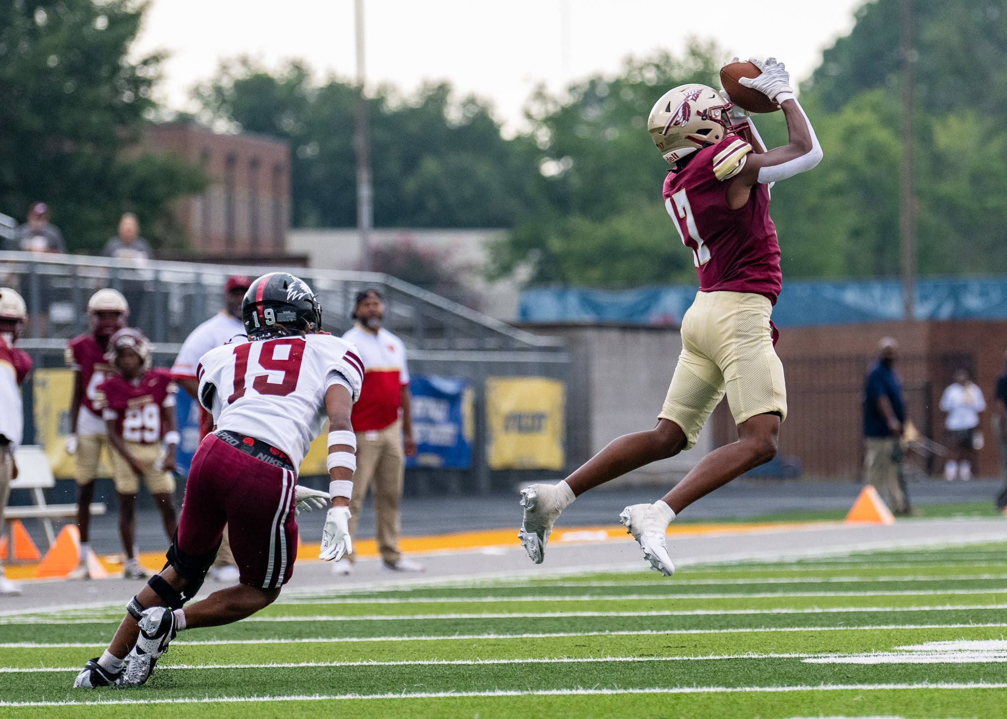 A football player wearing a garnet uniform with the number 17 catches a thrown ball while being defended by a player in an white uniform with the number 19.
