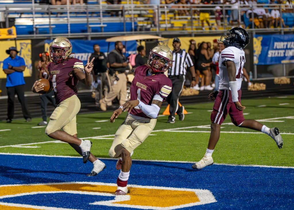 A player with a golden helmet emblazoned with a spear, a maroon jersey and gold pants holds the ball in one hand while making a peace sign in the other as he scores. A teammate in the same uniform and a defender, wearing a white jersey with maroon pants and a black helmet, follow.
