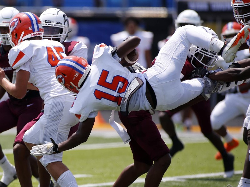 A football player in an all-white uniform with orange numbers, an orange helmet and white gloves is flipped parallel to the ground on a play while holding the ball.