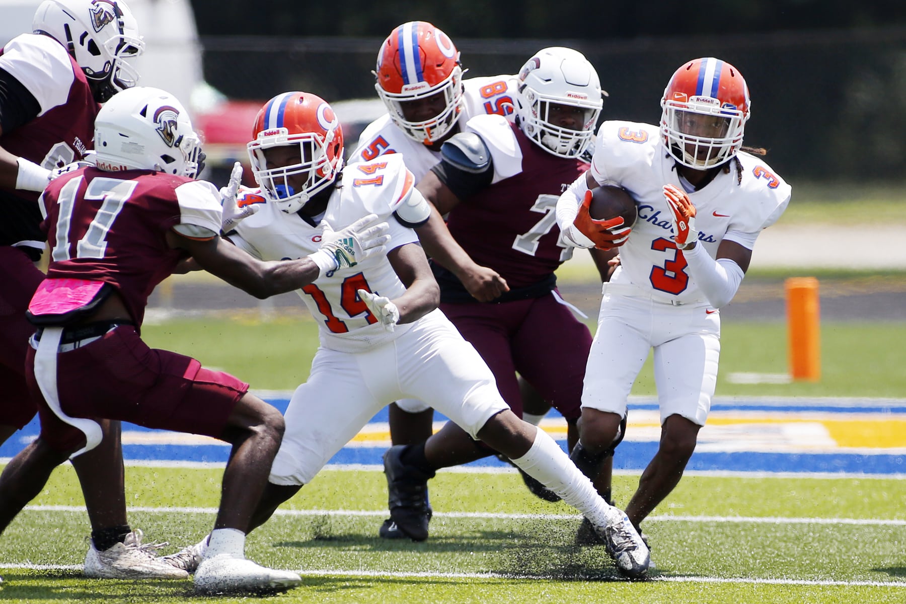 A football player in a white uniform with the number 5 runs with the ball between teammates and players in dark red uniforms.