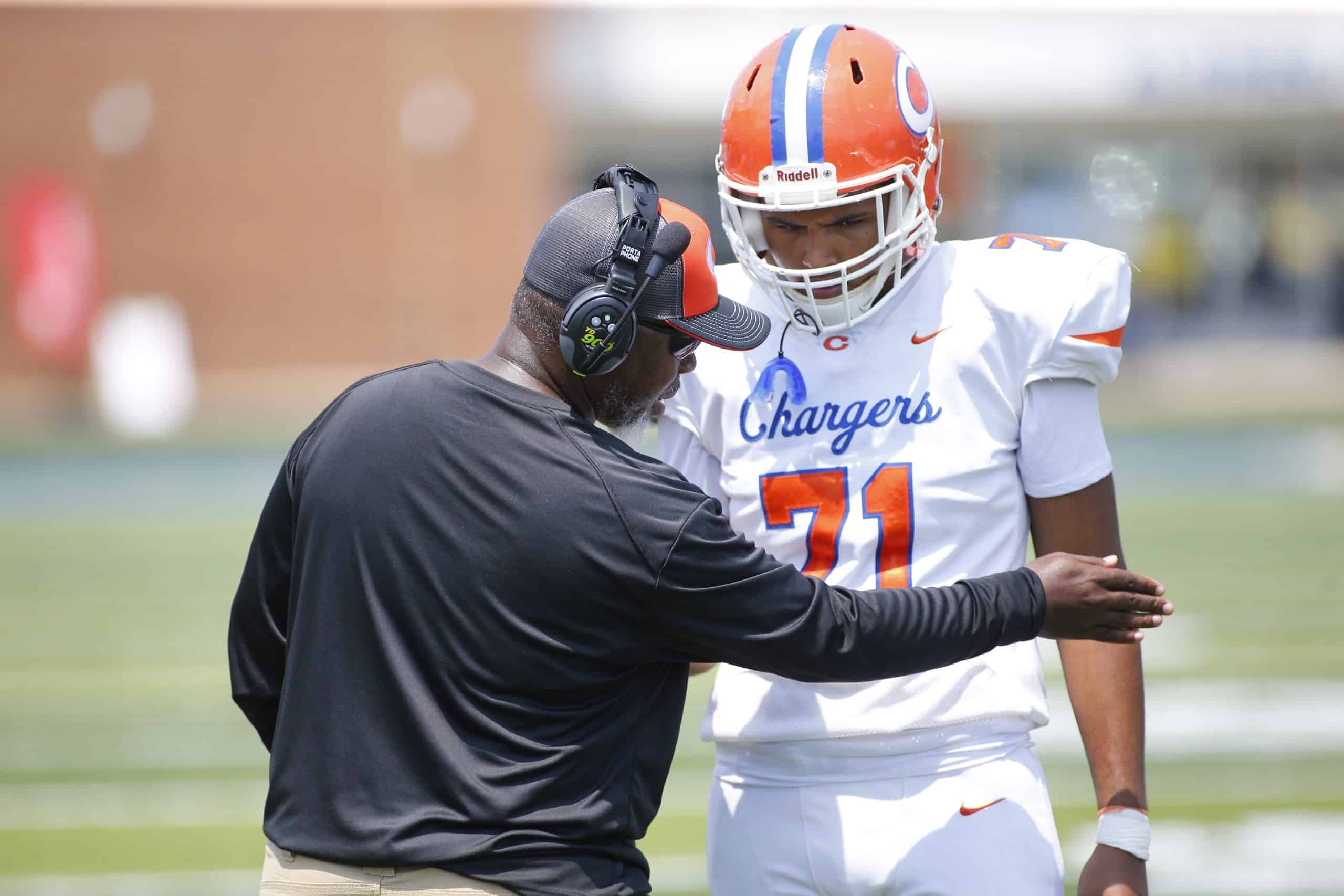 A football coach points to the right while taking with a player in a white uniform with the number 71.