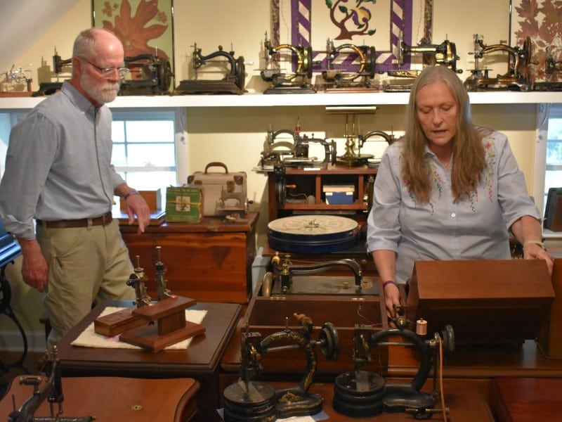 A man wearing a blue button-up shirt and khakis and a woman in a blouse inspect an old sewing machine in a room full of sewing paraphernalia.