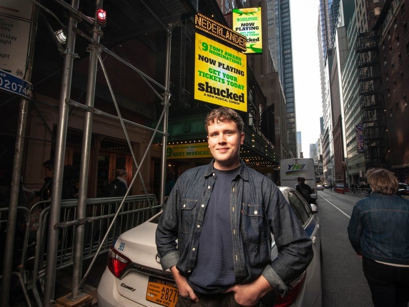 A man stands smiling on a city street near the entrance of a theater. Behind him, a bright yellow marquee reads "NOW PLAYING" and advertises a musical comedy titled "Shucked," which has received multiple Tony Award nominations. The theater's name, "Nederlander," is visible at the top of the marquee. The surrounding area shows tall buildings and a few people walking by.