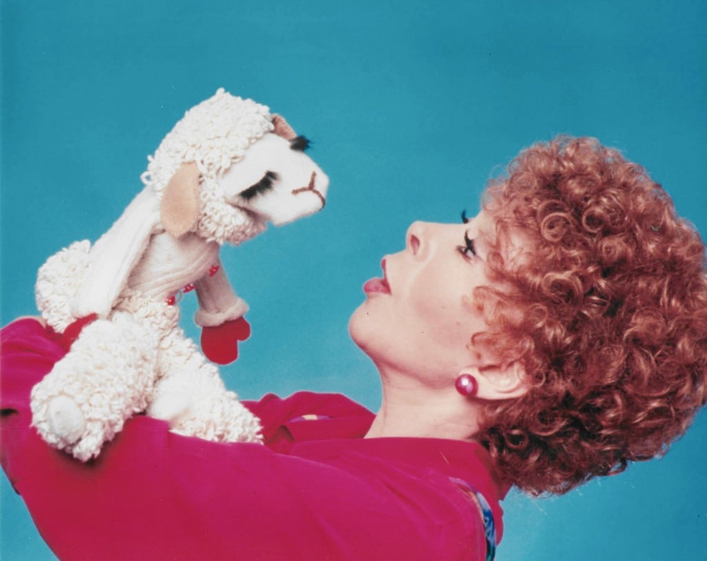 A woman with red curly hair holds up a sheep doll in front of her face, leaning back