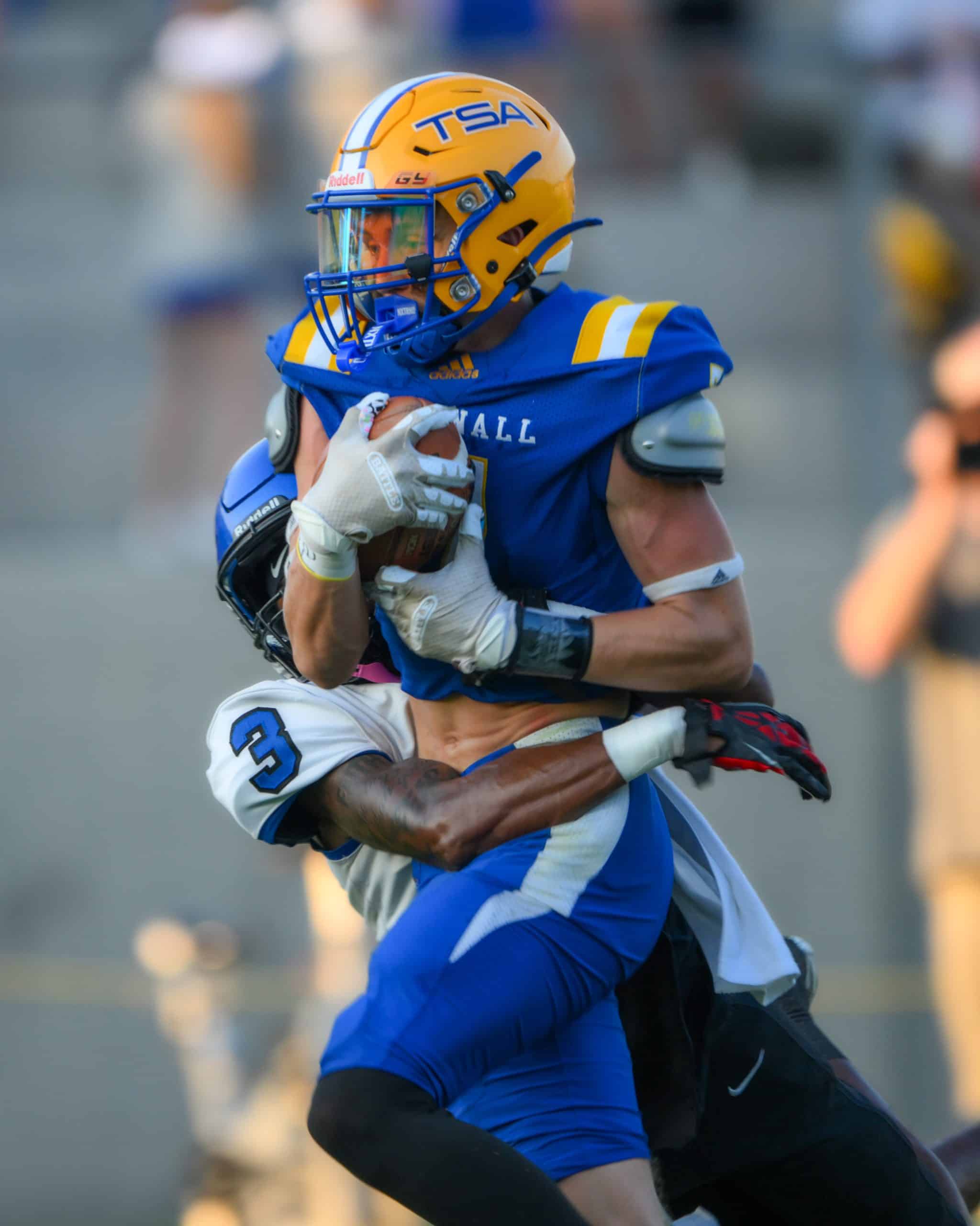 A football player in a blue uniform with the ball is tackled by a player in white uniform with the number 3.