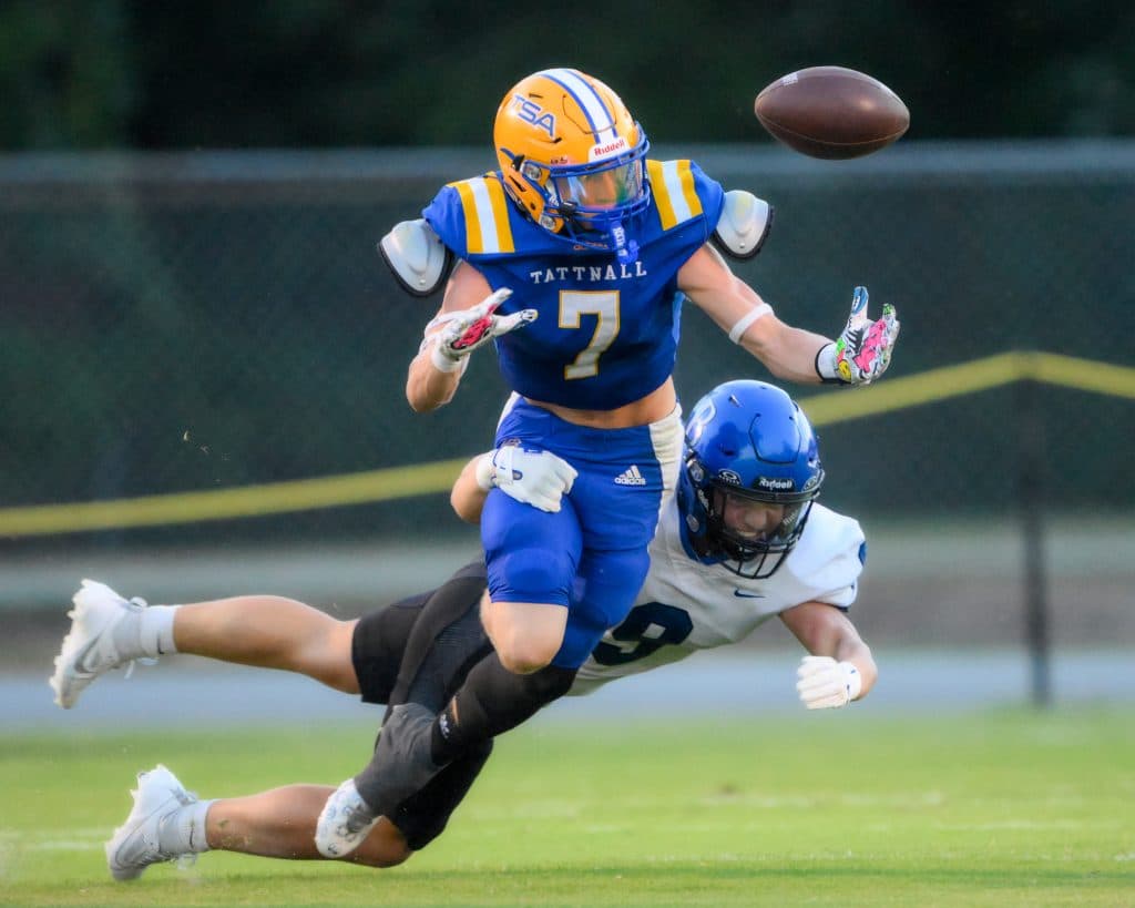 A player in an all-blue uniform with a yellow helmet that reads "TSA" grasps for a ball in mid-air as another player with a white jersey and blue helmet attempts to tackle him.