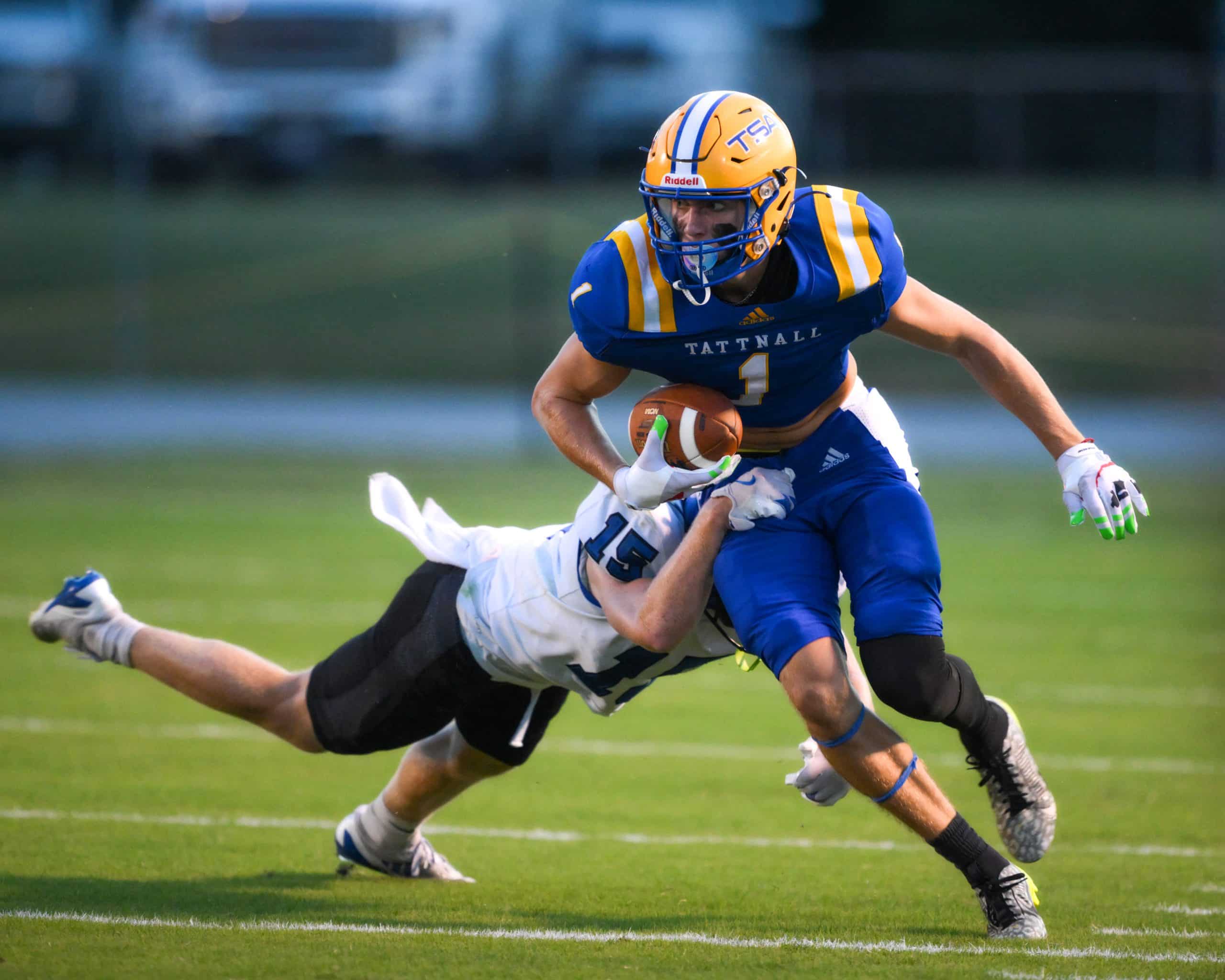 A football player in a blue uniform with the number 1 runs the ball while being tackled by a player in white uniform.