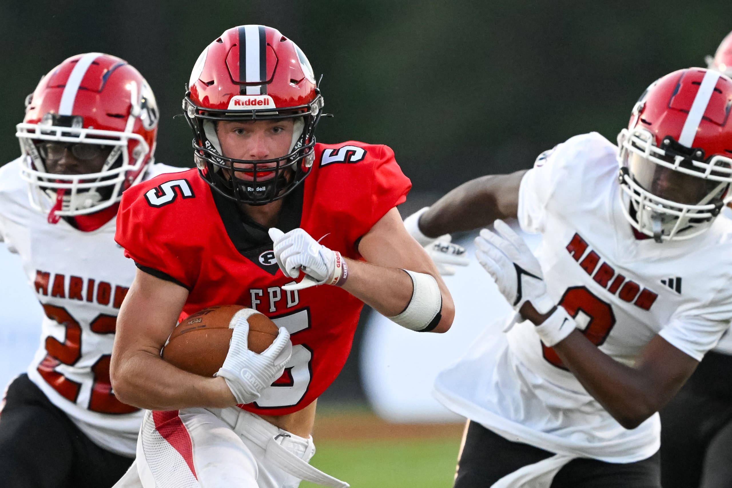 A football player in a red uniform and helmet with the number 5 is running with the ball, closely pursued by two defenders in white uniforms with red helmets. The defenders' jerseys display "Marion" in bold letters.