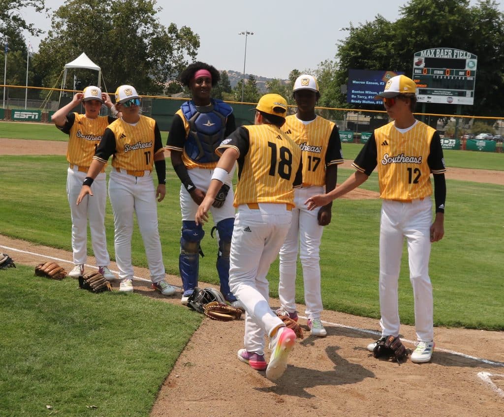 A player wearing a yellow jersey with black sleeves, a yellow baseball cap and white pants jogs past a line of teammates, low-fiving them.