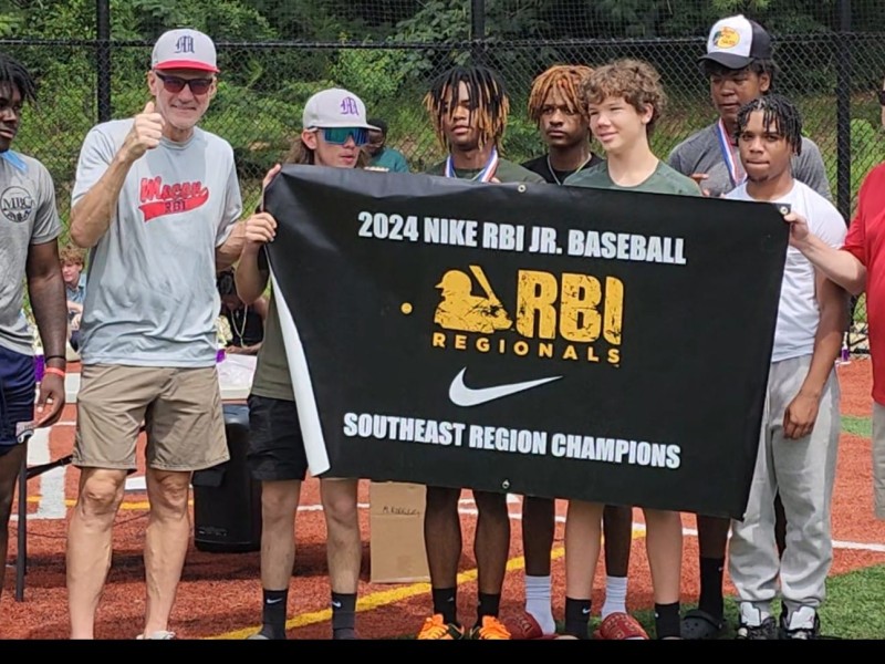 A group of players and coaches wearing various types of athletic wear surround a black banner with gold text showing a logo reading Nike RBI.