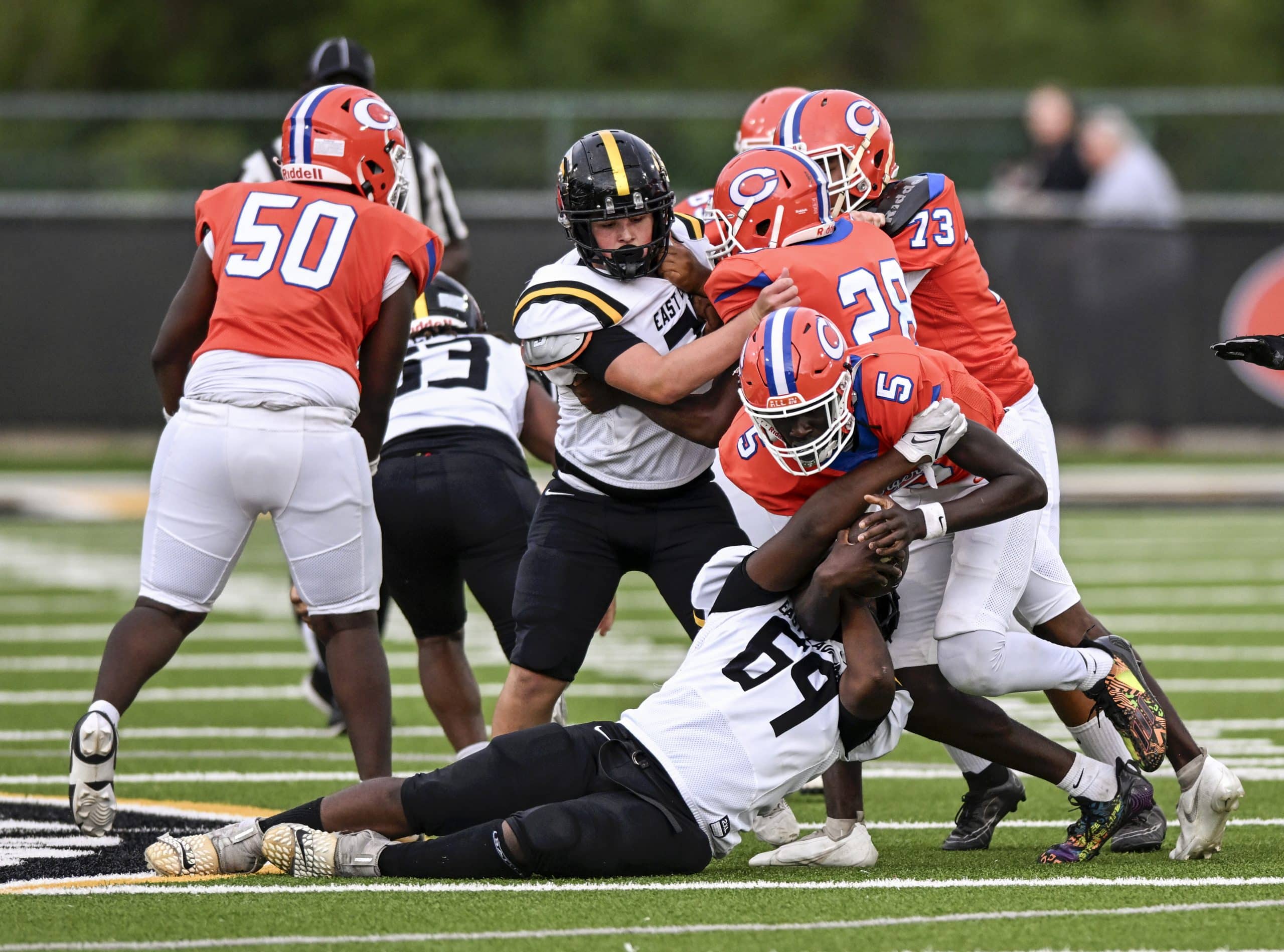 A group of football players in action on the field. Players in white jerseys and black pants are tackling a player in a orange and white uniform. One player in an orange jersey, number 5, is being taken down a player in a black helmet. The scene is intense, with players struggling for control of the ball.
