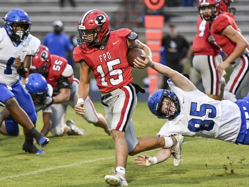 A football player in a red jersey and helmet, number 15, runs with the ball while a player in a white jersey and blue helmet, number 85, attempts to tackle him by grabbing his arm. Other players in blue and red jerseys are in pursuit or engaged in tackles in the background.