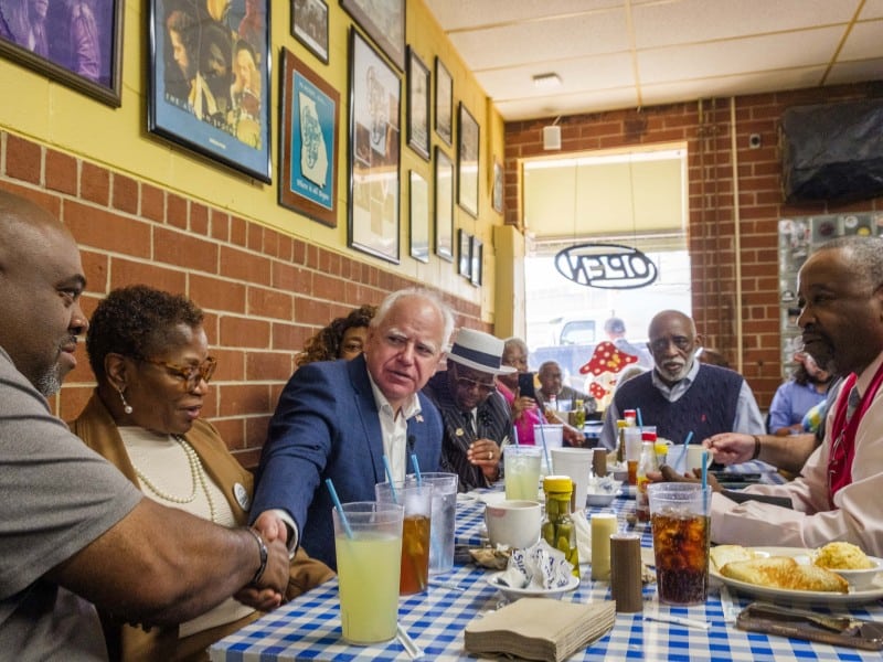 People sit around a booth with a blue and white checkered table cloth. One man shakes another man's hand.