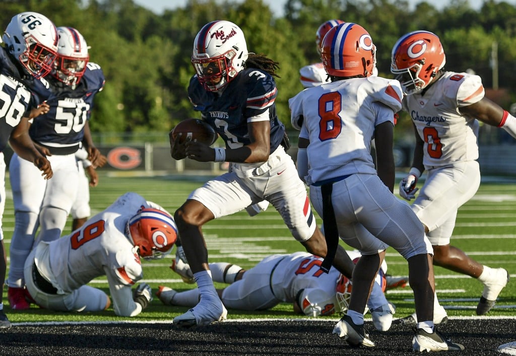 A football player in a navy blue uniform with white pants, number 3, runs with the ball while navigating through defenders in orange and white uniforms. Players from both teams are scattered on the ground, with one wearing number 9 on the ground in front of the runner. Two defenders in white and orange, wearing numbers 8 and 0, are preparing to block or tackle him. The scene is set on a sunny day on a green football field.