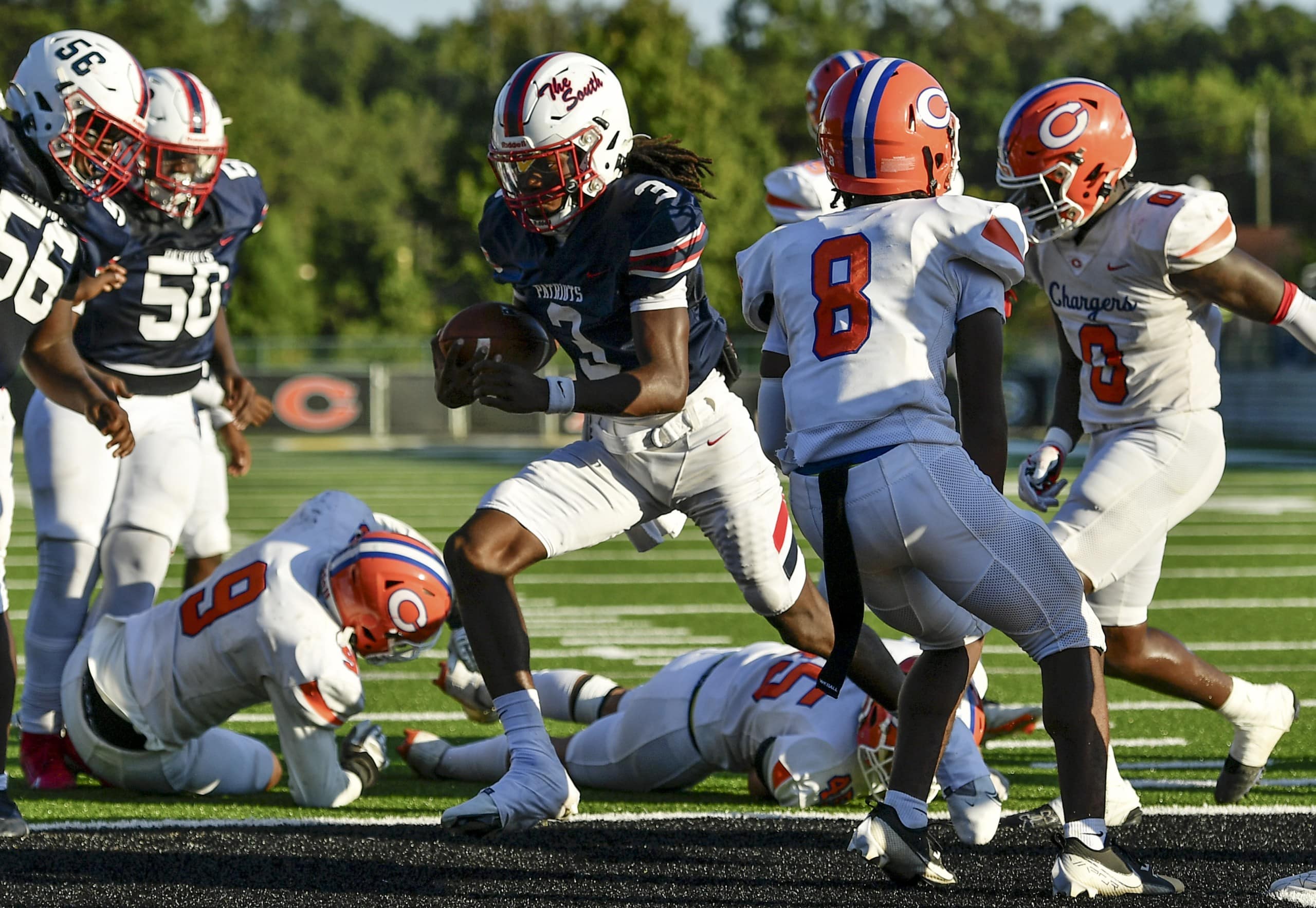 A football player in a navy blue uniform with white pants, number 3, runs with the ball while navigating through defenders in orange and white uniforms. Players from both teams are scattered on the ground, with one wearing number 9 on the ground in front of the runner. Two defenders in white and orange, wearing numbers 8 and 0, are preparing to block or tackle him. The scene is set on a sunny day on a green football field.