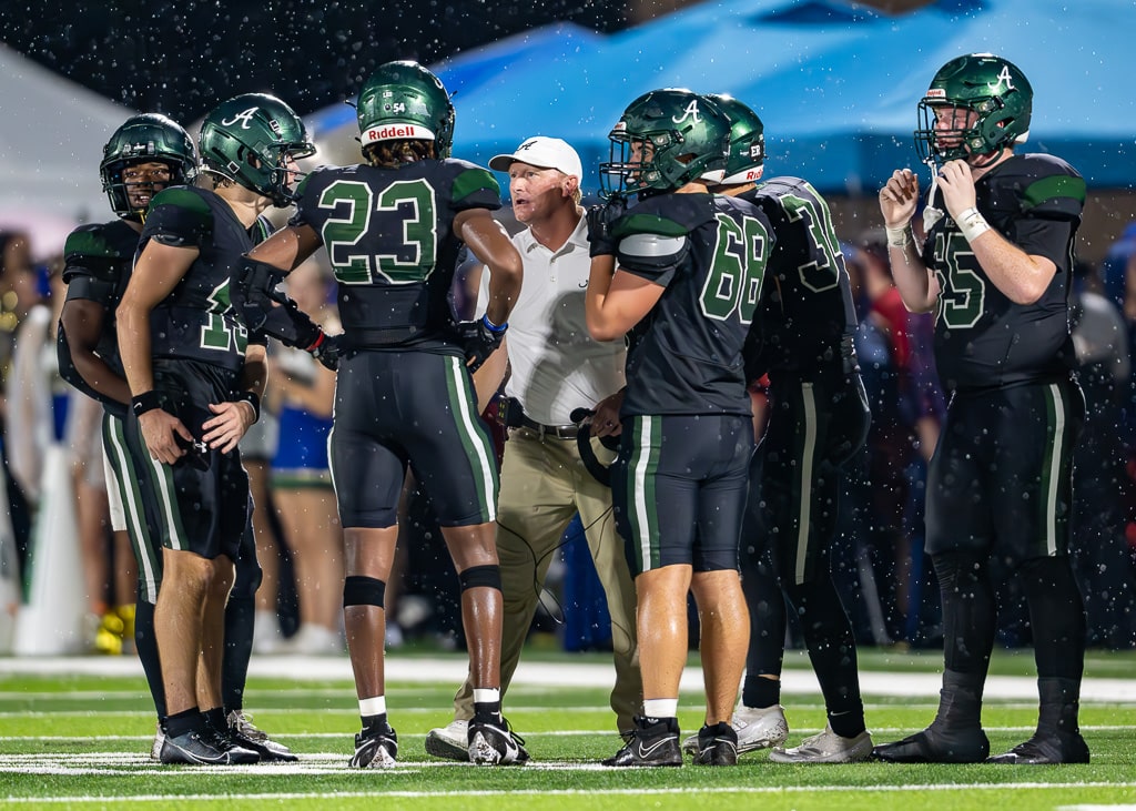 A group of football players in black uniforms with green helmets gather around their coach on the field during a game. The coach, wearing a white shirt and hat, is speaking to the players, with number 23 standing in the center. Light rain is falling, and the scene takes place under stadium lights, with spectators and tents visible in the background.