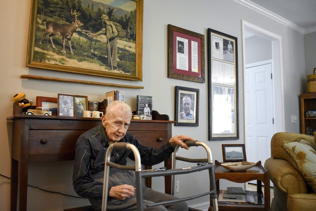 An elderly man sits in a room using a walker, with framed photos and articles on the wall behind him. A large painting of a man and a deer is displayed above a wooden desk, which holds family photos and memorabilia.