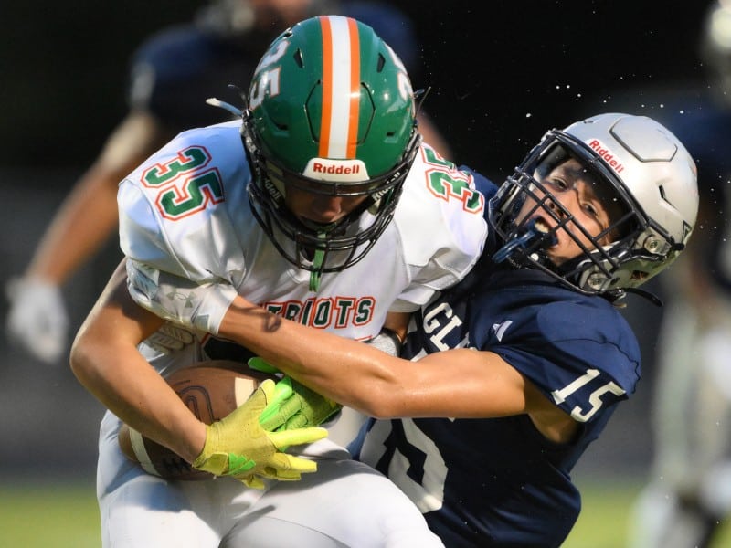 A football player in a white uniform with the number 35 and a green helmet is tightly holding the ball while being tackled by a player in a navy blue uniform with the number 15. The scene captures a close-up, intense moment of contact during the game.