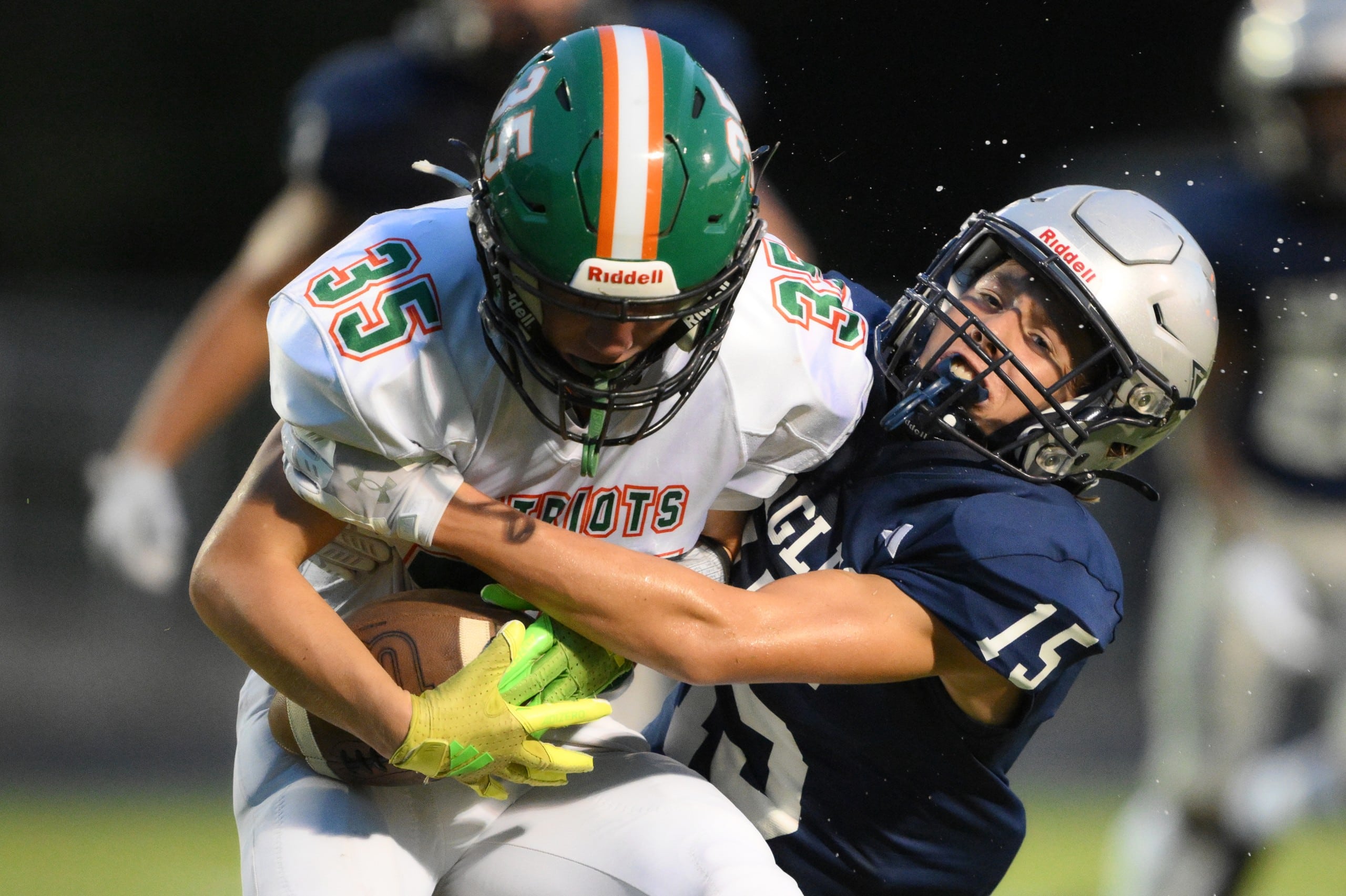 A football player in a white uniform with the number 35 and a green helmet is tightly holding the ball while being tackled by a player in a navy blue uniform with the number 15. The scene captures a close-up, intense moment of contact during the game.