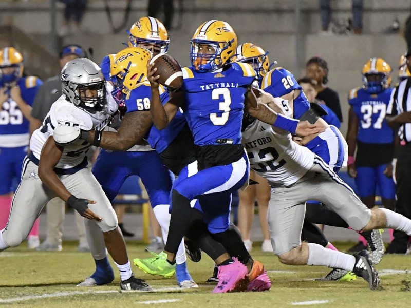 A football player in a blue and yellow jersey, holding the ball, is being tackled by two opposing players in white and gray uniforms. The quarterback is attempting to evade the defenders, with one player grabbing his arm and another reaching for him. Several teammates and a referee are visible in the background. The action takes place on a well-lit field.
