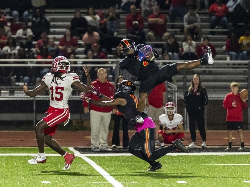 A football player in a black uniform and helmet jumps high to catch a pass, holding the ball securely while extending his legs in the air. Another player in black attempts to block a nearby opponent in a white and red uniform with the number 15. Spectators in the stands watch the action under bright field lights.