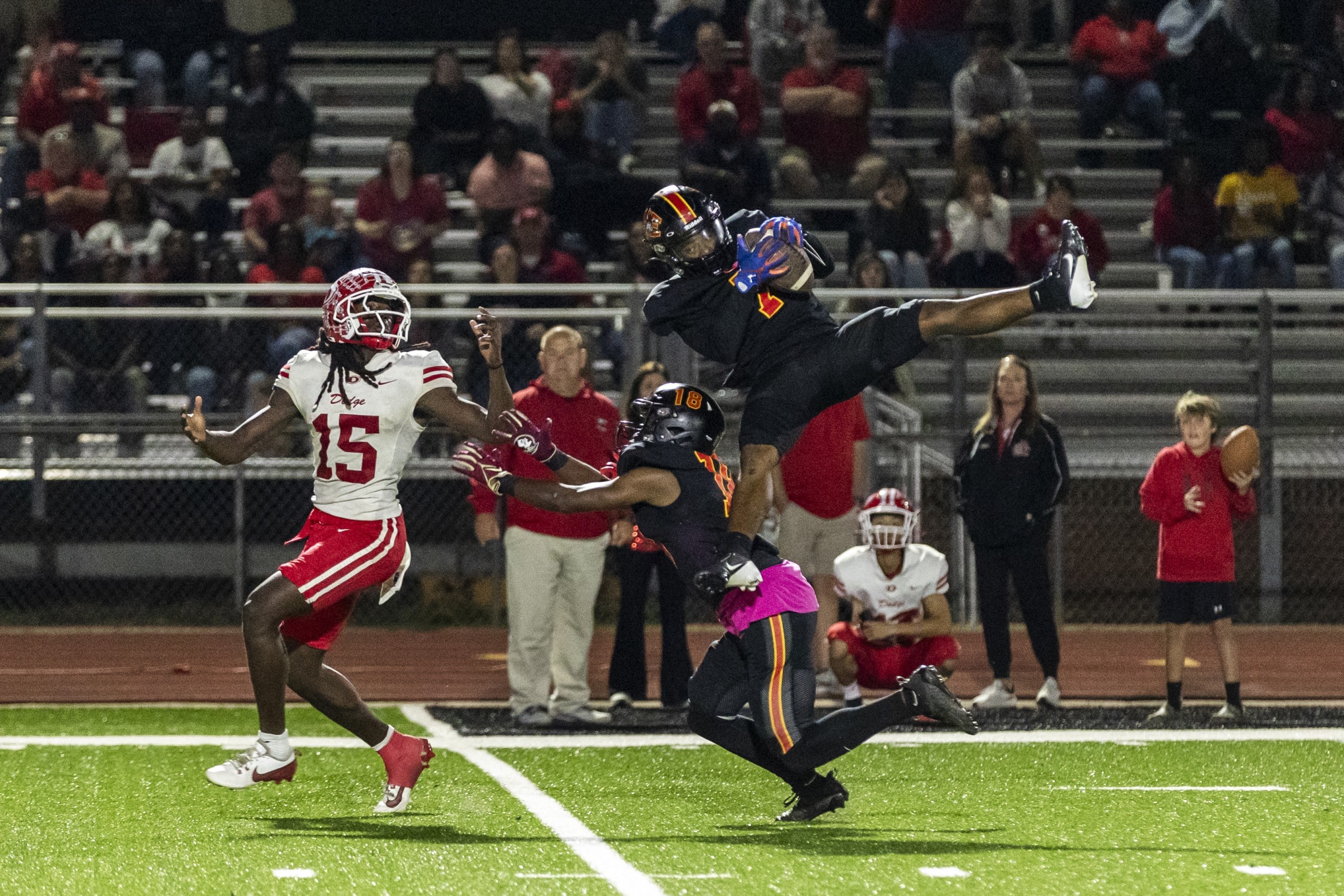 A football player in a black uniform and helmet jumps high to catch a pass, holding the ball securely while extending his legs in the air. Another player in black attempts to block a nearby opponent in a white and red uniform with the number 15. Spectators in the stands watch the action under bright field lights.