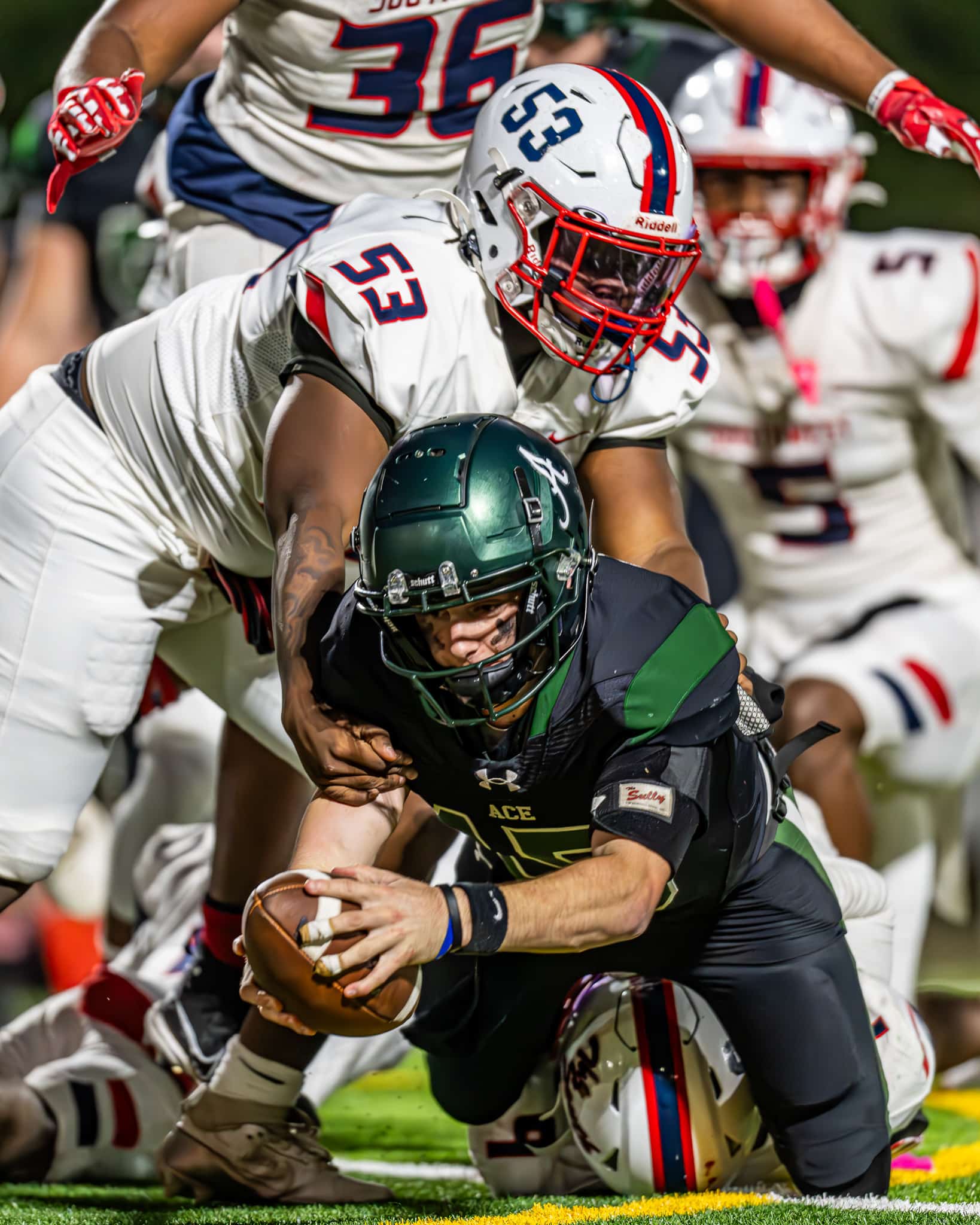 A football player in a dark green and black uniform is being tackled while reaching forward with the ball, likely attempting to score or gain yardage. A defender in a white uniform with red and blue accents grabs him from behind. Several other players are in the background, adding to the intensity of the scene. The action is happening on a well-lit field.