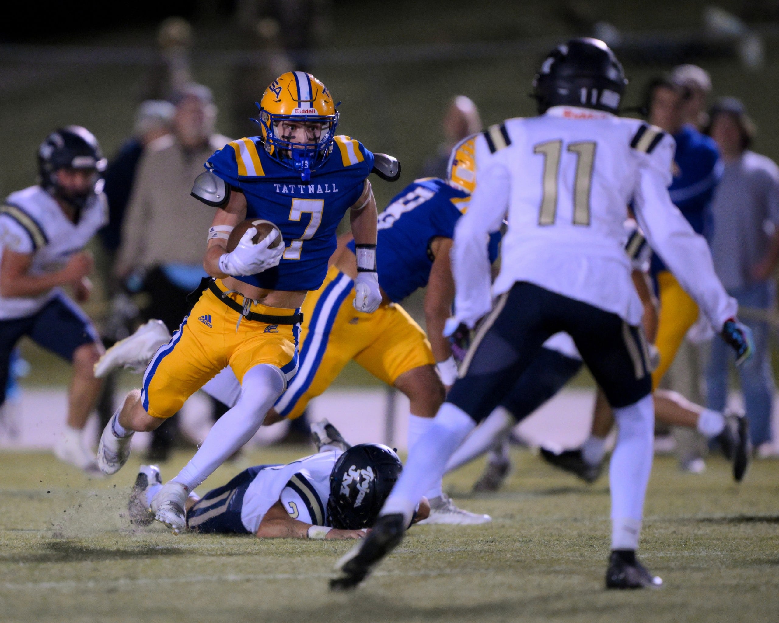A football player in a blue and yellow Tattnall uniform runs with the ball, evading a defender who is on the ground after a missed tackle. Another defender in a white and black uniform approaches from the front. The action is taking place on a grassy field under stadium lights.