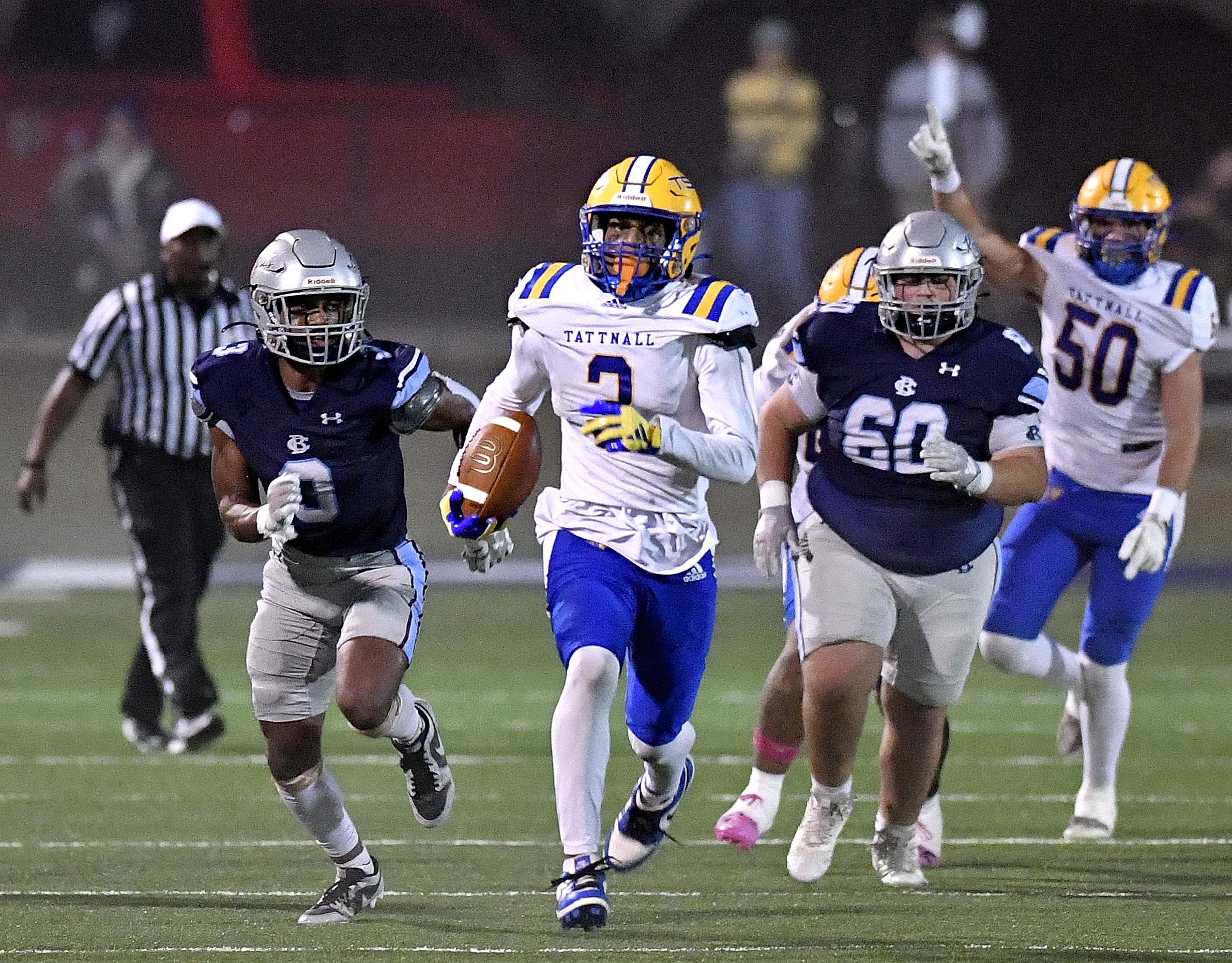 A football player in a white and blue uniform with the number 2 runs with the ball, pursued by two players in dark blue uniforms. Teammates and opponents follow close behind, with one player raising a hand, possibly signaling. The scene takes place under bright lights on a green field.
