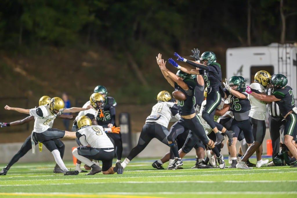 A football kicker in a white and gold uniform attempts a field goal, with his teammate holding the ball in position. Opposing players in black uniforms jump and reach high, trying to block the kick. Other players clash on the line of scrimmage, focusing on their blocks. The play takes place under bright stadium lights on a green turf field.