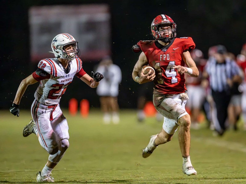 A football player in a red uniform with "FPD" and number 14 sprints forward with the ball, pursued by an opponent in a white "Patriots" uniform with number 22. The player in red has a determined expression and appears to be dodging the defender. The field is illuminated, and other players and spectators are blurred in the background.