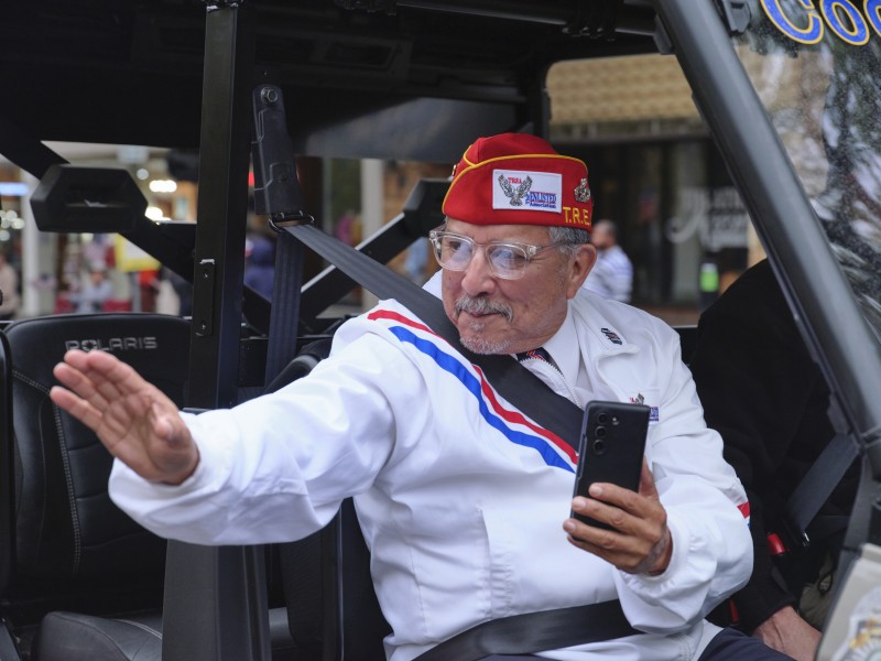 An elderly man in a white uniform with red, white, and blue detailing and a red veteran’s cap sits in a vehicle, smiling and waving to people outside. He holds a smartphone in his other hand. The background shows a street scene with blurred buildings and pedestrians.